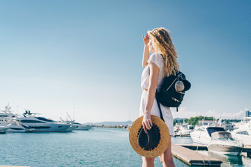 having made from a palm a visor above the eyes from the sun, a curly blonde looks into the distance while standing on the pier. she has a backpack behind her - 315867729