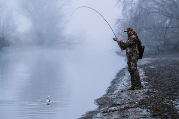 Fisher man fishing with spinning rod on a river bank at misty foggy winter, spin fishing, prey...
