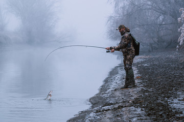 Fisher man fishing with spinning rod on a river bank at misty foggy winter, spin fishing, prey...