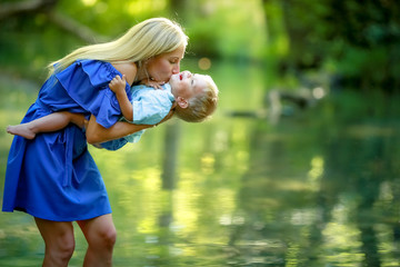 A mother in a bright summer dress stands in a river or lake in the forest and plays with a baby in her arms.