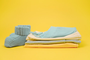 Cute blue booties for newborn baby boy and heap of clothes. Clothes for infant on yellow background. Expecting a baby concept. - Powered by Adobe