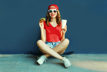 Cool young woman with burger and coffee cup sitting on skateboard wearing baseball cap, sunglasses over blue wall background © rohappy