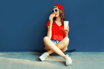 Cool young woman with burger and coffee cup sitting on skateboard wearing baseball cap, sunglasses over blue wall background © rohappy