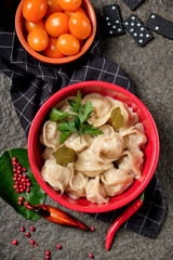 Russian pelmeni meat with butter, chili pepper, branch of cherry tomatoes, fresh vegetables, herbs on black stone background. Bowl with tasty dumplings and bay leaf. Rustic style.