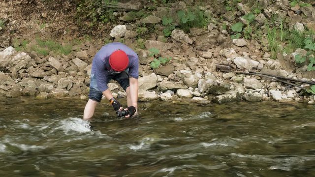 Man with a camera standing in a rocky river and taking pictures of the water surface. Stock footage. Male hiker with a camera shooting videos of the forest stream.