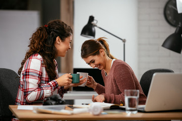 Two businesswomen having fun in office. Beautiful colleagues drinking coffee at work.