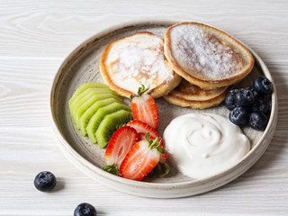 Delicious breakfast - fritters pancakes, sprinkled with powdered sugar with fruits and berries and sour cream on plate on a wood table.