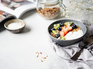 Delicious breakfast - the cup of coffee, dry coconut in bowl, granola with yogurt and berries and open magazine on the table. Copy space.