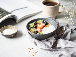 Delicious breakfast - the cup of coffee, dry coconut in bowl, granola with yogurt and berries and open magazine on the table. Copy space.