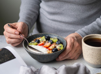 Uncertain girl in grey dress at the table with breakfast. Bowl with yogurt, granola and berries and cup of black coffee and open magazine on grey table
