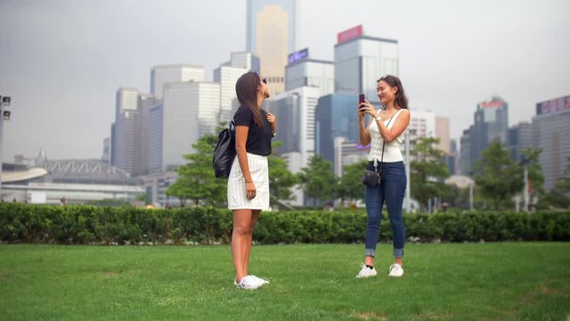 Two Young Women Take Photos in the Park with Green Grass and Buildings Around