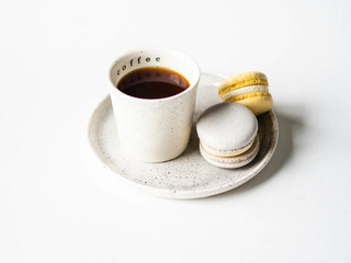 Two pastel macaroons with white ceramic cup of black coffee on ceramic plate on white background