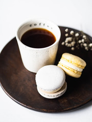 Two pastel macaroons with white ceramic cup of black coffee on wood plate on white background
