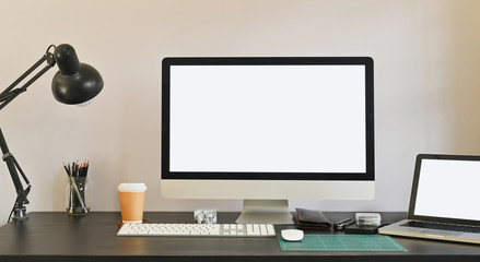 Photo of white blank screen computer and white blank screen laptop putting on the table together including lamp, coffee cup, mouse pencil holder and personal equipment. Modern working desk concept.