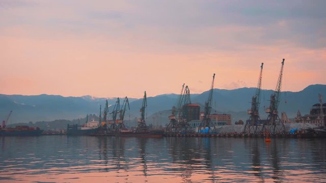 Stunning view of the sea port of Batumi with cranes, barges and ships in the sea against the backdrop of mountains at sunset.