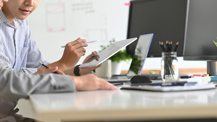 Close-up image of young businessman in white striped suit holding a white blank screen tablet and pointing/showing/discussing/brainstorming/planning to his young creative colleague.