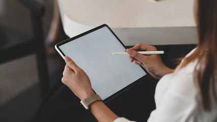 Close-up image of confident woman hand holding black tablet with white blank screen display and using pencil for writing or drawing on it. Behind shot.