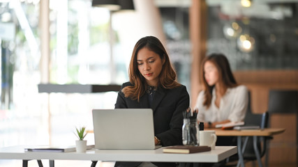Photo of young business woman in look good black suit typing on her white laptop while sitting at work desk in the modern office. Sitting over colleague background.