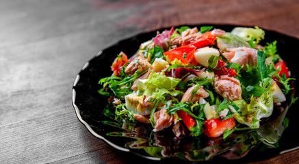 Fresh salad with fish, arugula, eggs,red pepper, lettuce, fresh sald leaves and tomato on a black plate on wooden table background