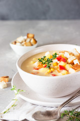 Bowl of homemade corn chowder soup with potatoes, carrots, red bell pepper and croutons on a light grey stone background. Delicious cozy first course, comfort food.