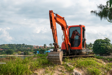 Small orange excavator on a ground against blue sky and sea for a works on construction site. Small...
