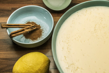 Creamy rice pudding or rice pudding in a large bowl next to a whole yellow lemon, a bowl with ground cinnamon and cinnamon stick and a bowl with sugar.Only brown walnut background.