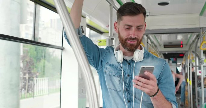 Handsome Bearded Man Standing at the public transport, texting Messages on Mobile Phone. Holding Handrail with his right Hand. Search for some Songs to listen with his Big Headphones. Looking Happy