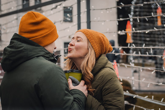 Attractive young couple in love, hugging and kissing on the street on the background of garlands of lights. Outdoor portrait of stylish fashion couple with coffee. Valentines day, christmas concept.