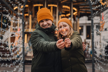 Beautigul couple celebrates together at the christmas market. Girl and guy with a sparkles in their hands wearing orange hats and green jackets. Winter holidays concept.
