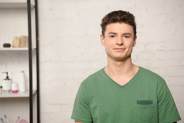 Obraz premium handsome young man in green t-shirt smiling at camera while standing near rack with cosmetics