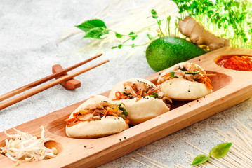 Bao - steamed pie with meat filling and fresh vegetables. Bao on beautiful wooden plate with chinese chopsticks, on light concrete background, decorated with fresh vegetables. Traditional chinese food