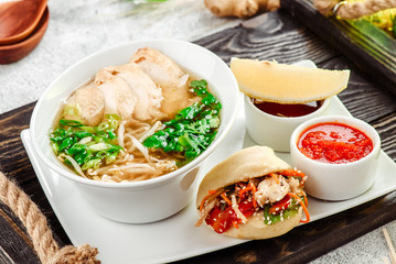Asian noodle soup, ramen with chicken, tofu, vegetables in white plate on a dark restik tray in beautiful composition with fresh herbs on light concrete background. Close up. Top view.