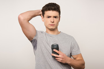 displeased man in grey t-shirt with sweaty underarm holding deodorant isolated on grey