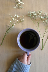 Unrecognizable person in a cozy sweater holding a cup of tea, with gypsophila flowers on wooden table. Flat lay. 