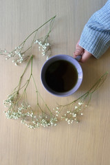 Fototapeta na wymiar Unrecognizable person in a cozy sweater holding a cup of tea, with gypsophila flowers on wooden table. Flat lay. 