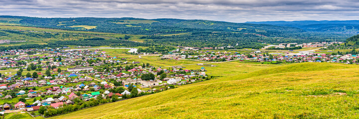Fototapeta na wymiar Panoramic view from hill to plain with small town in summer cloudy day. Picturesque urban landscape with many colorful houses. Sim, Chelyabinsk region, Russia. Travel blog concept