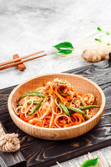 Pan-Asian food. Noodles with vegetables and asparagus beans in beautiful wooden plate on dark rustic tray on light concrete background. Beautiful composition. Close-up. Space