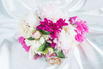 Valentine's day or Wedding concept. Two wedding rings and bouquet of pink and purple peonies on white satin background