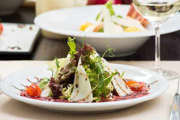Fresh salad with beef carpaccio, parmesan, tomatoes and sauce on a white plate and a glass of white wine on a table in a restaurant. Close-up. Space
