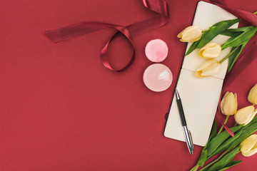 top view of tulips with silk ribbon, macarons and empty notebook with pen isolated on red