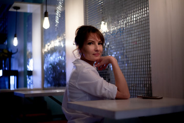 Adult beautiful woman sit in restaurant and waiting the order in evening. The female person wearing white shirt.