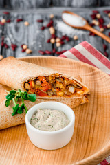Burrito with vegetables, decorated with soy sprouts, served with cream sauce, on wooden plate on gray wooden background