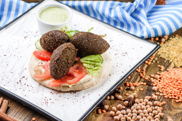 National Israeli dish - Pita with falafel, lettuce, tomato slices and sauce on a white square plate, on a dark wooden background decorated with legumes and with a blue napkin. Close up. Space