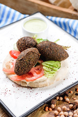 National Israeli dish - Pita with falafel, lettuce, tomato slices and sauce on a white square plate, on a dark wooden background decorated with legumes and with a blue napkin. Close up. Space