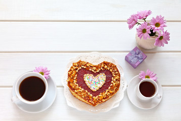 Top view on festive table with a heart-shaped homemade cake, two cups of coffee and a flowers. Beautiful congratulations on Valentine's Day or Happy Birthday. Flat lay, copy space, close-op, top view