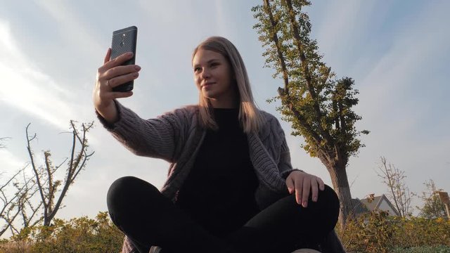 Young attractive blonde woman sitting in the lotus position doing selfie outdoor in park