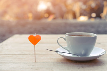A red heart shaped candle with a cup of coffee and a coffee spoon on a saucer placed on a wooden table in the garden. Valentine day and love concept.