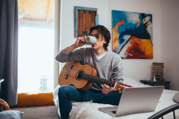 teenager playing guitar in his room at home, using laptop computer for online lessons