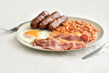 English breakfast with bacon, egg, baked beans and beef sausages on a white plate with a fork and a knife on the table. Selective focus.