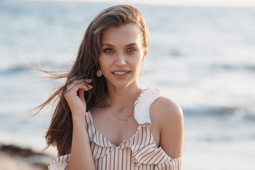 Beautiful young woman posing against the sea and skyline.Happy woman relaxing near the sea on beach.Hipster girl in beige dress standing and tanning on beach near sea with waves,sunny warm weather.